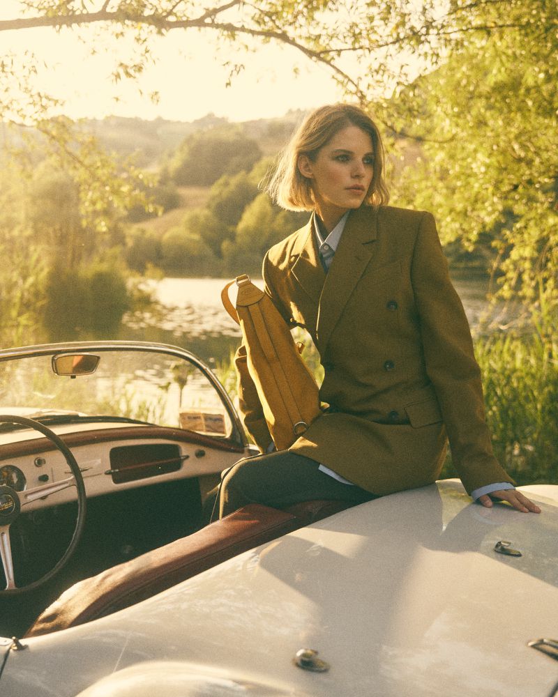 Blonde woman in olive blazer sitting on the hood of a white convertible car, with a river and trees in the background, outdoors.
