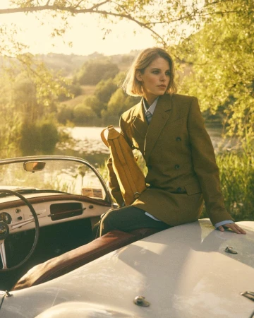 Blonde woman in olive blazer sitting on the hood of a white convertible car, with a river and trees in the background, outdoors.