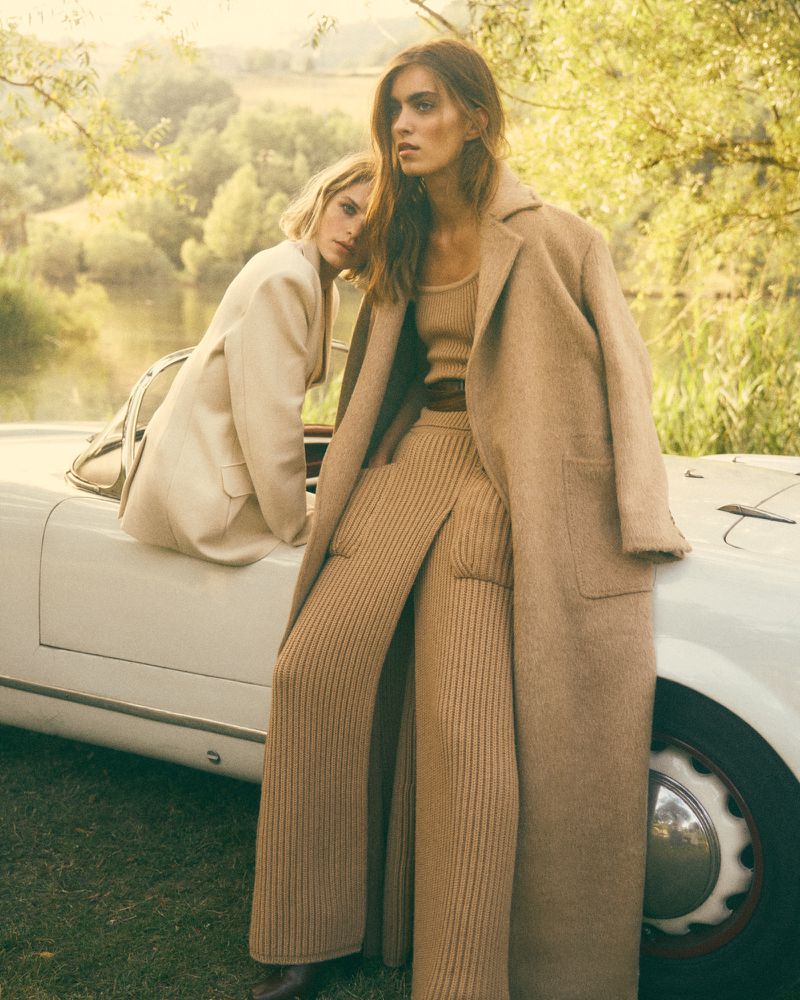 Two women in tan-colored outfits pose on a classic white car in a sunlit outdoor setting with green grass and trees.