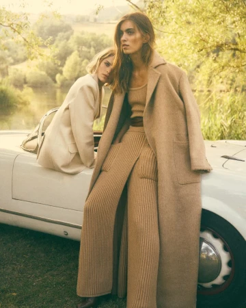 Two women in tan-colored outfits pose on a classic white car in a sunlit outdoor setting with green grass and trees.