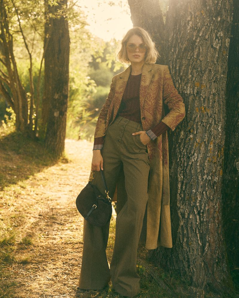 A woman in a patterned coat poses by a tree in a sun-drenched forest. She wears green flared pants and a dark handbag. Light flares.