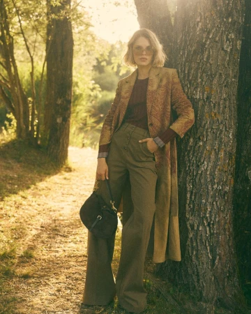 Model wearing a long patterned coat, wide-leg khaki trousers, and sunglasses leaning against a tree trunk in sunlit woods.