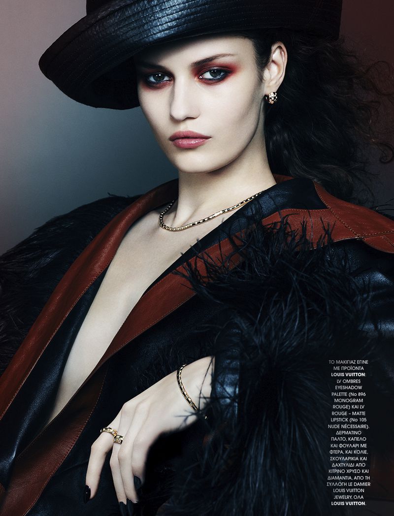 Portrait of a woman wearing a wide-brimmed hat and dark clothing, striking a pose in a studio setting with soft lighting and a makeup-heavy look.