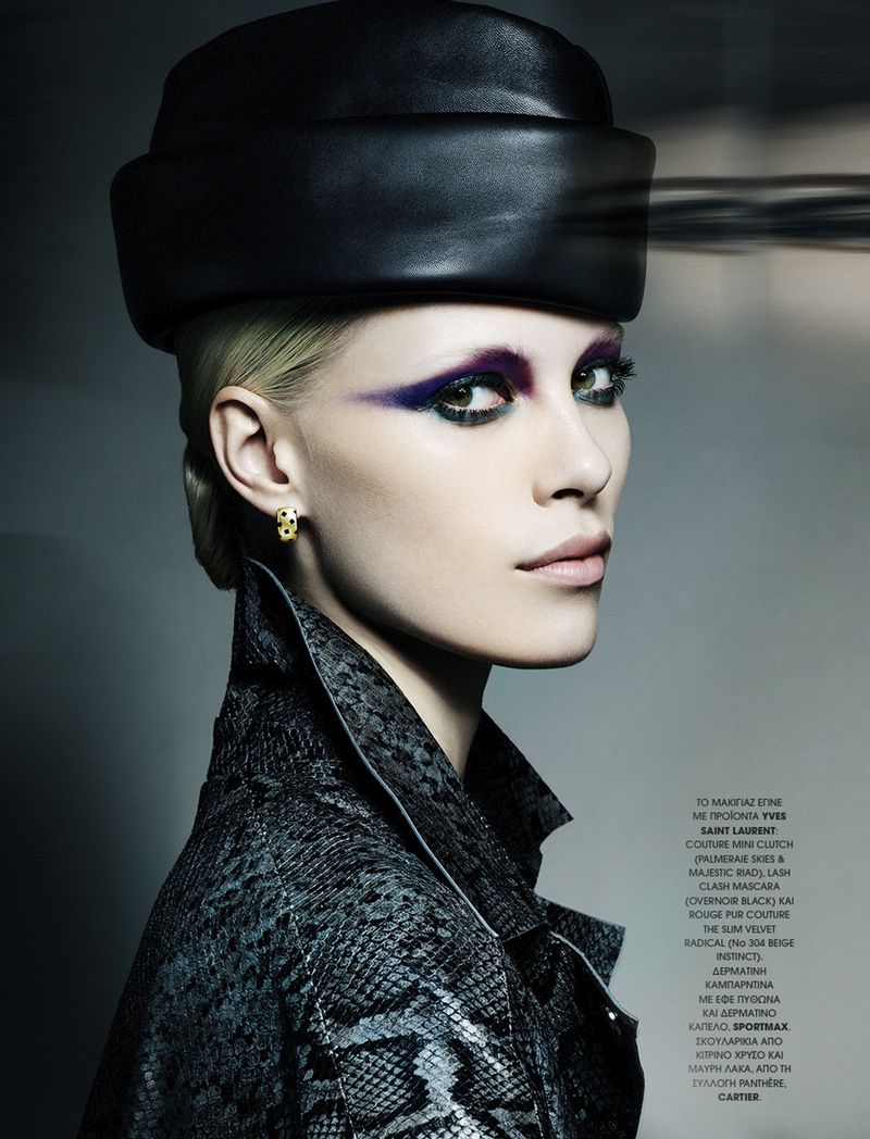 Fashion editorial portrait of a fair-skinned adult woman wearing a black hat and a snakeskin jacket against a neutral background.