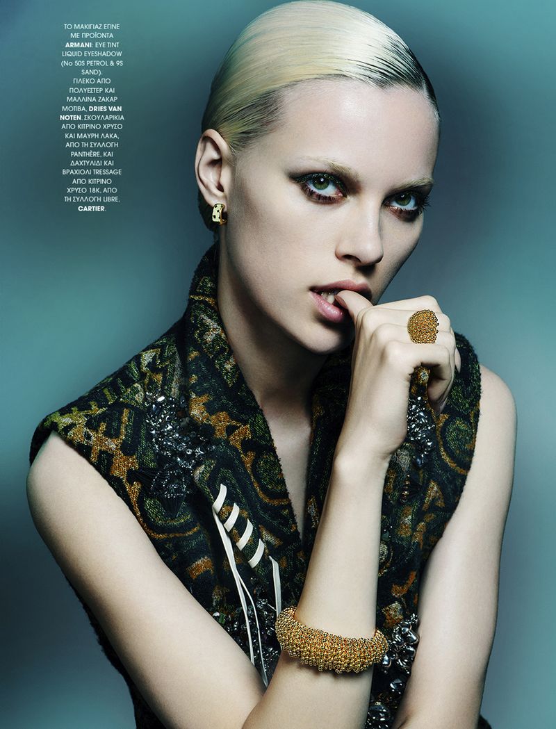 Fashion portrait of a blonde woman wearing a patterned vest and gold jewelry, posing against a teal backdrop with moody, studio lighting.