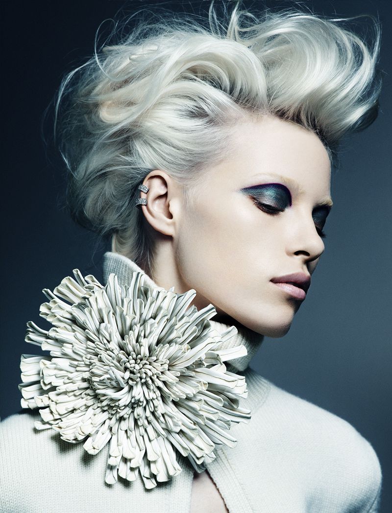Close-up of a woman with striking makeup, platinum blonde hair styled upwards, wearing a large, white floral collar or accessory.