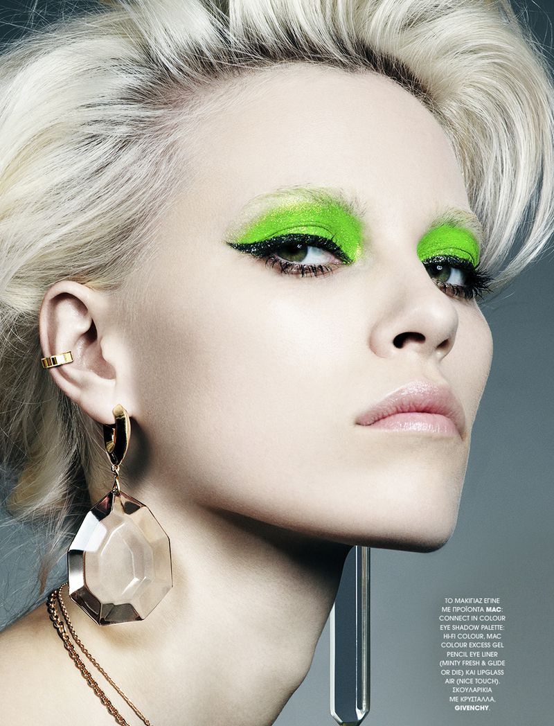 Close-up of a blonde woman with bright green eye makeup, gold jewelry, and pale skin, showcasing a fashion and beauty editorial shot.