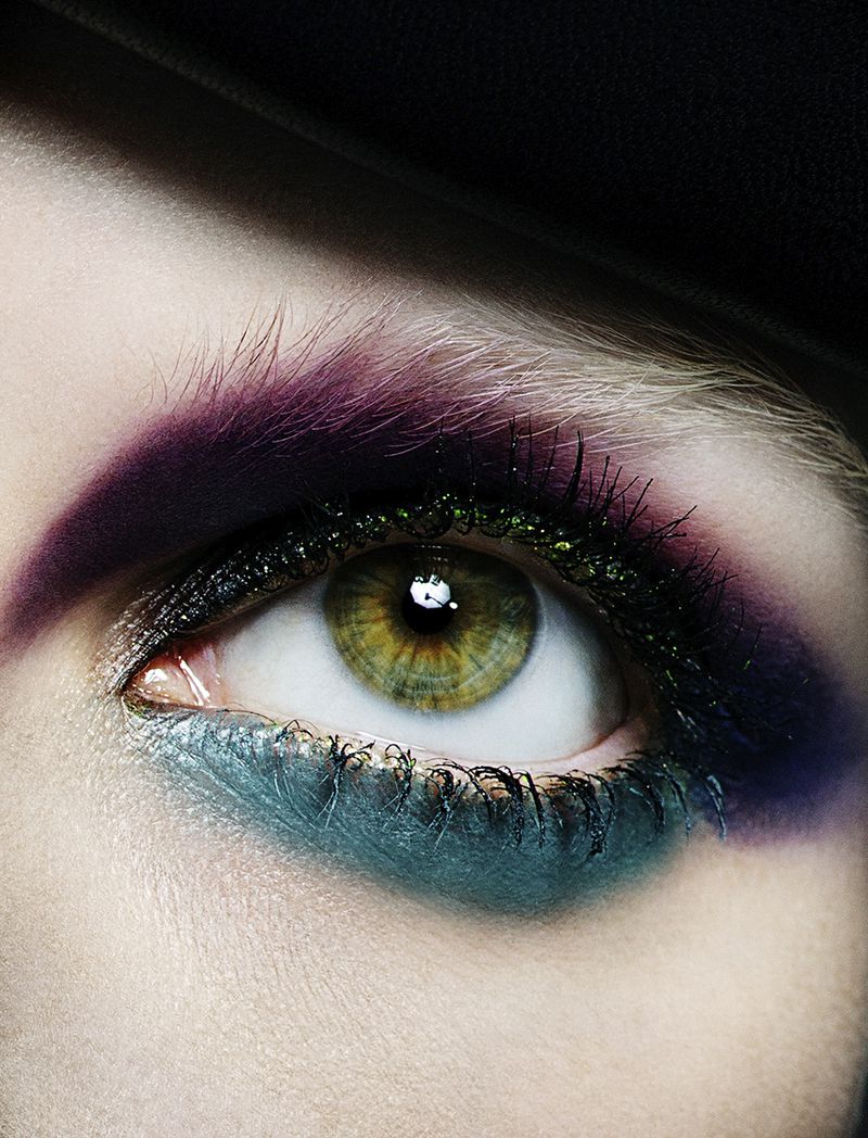 Close-up of a hazel eye with artistic makeup featuring purple, teal, and glitter eyeliner, set against a dark, neutral background.
