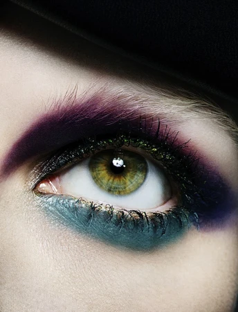 Close-up of a hazel eye with artistic makeup featuring purple, teal, and glitter eyeliner, set against a dark, neutral background.