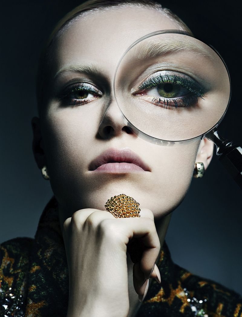 Close-up portrait of a fair-skinned woman with a magnifying glass over her eye, wearing a patterned jacket and large statement ring.