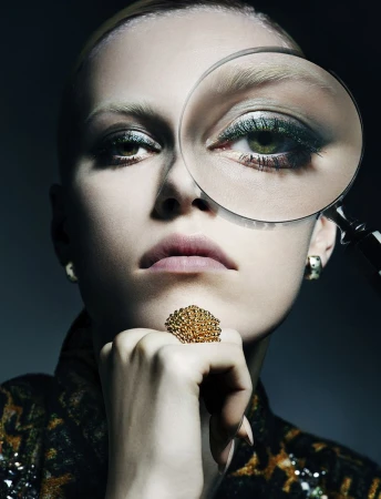 Close-up portrait of a fair-skinned woman with a magnifying glass over her eye, wearing a patterned jacket and large statement ring.