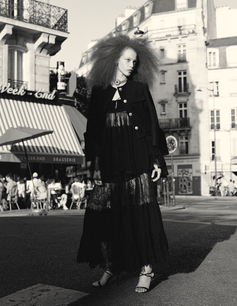 Fashion shot of a woman with large hair wearing a dark dress and coat, walking on a city street in front of a brasserie in monochrome.