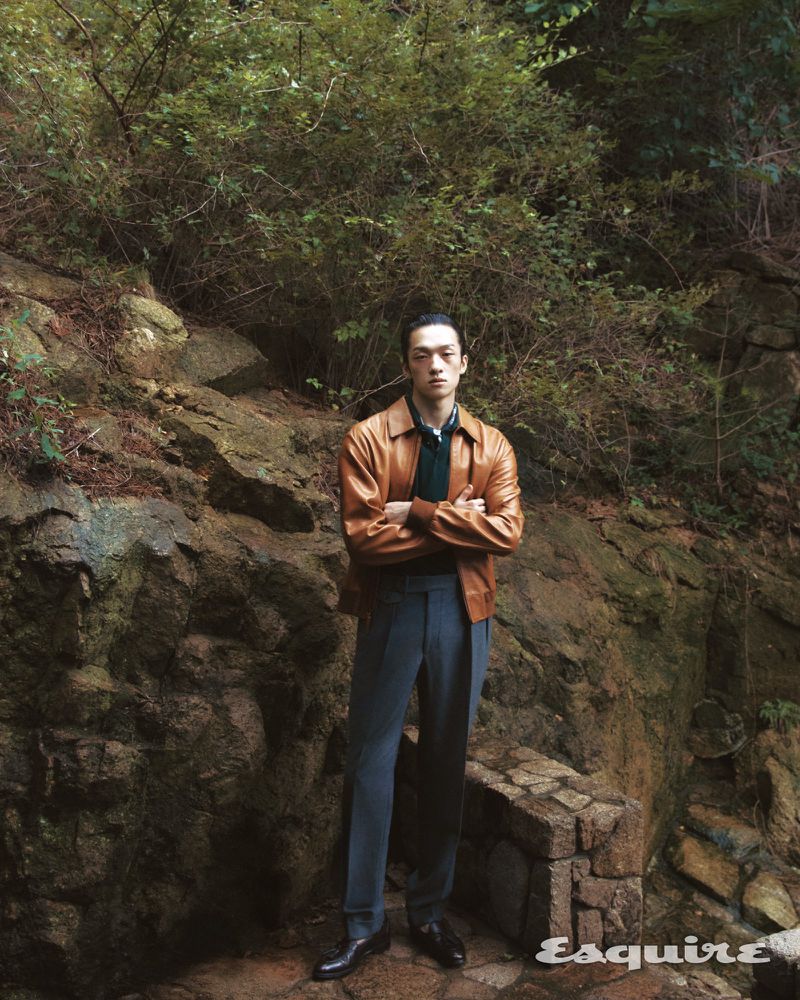 Stylish portrait of an adult Asian man in a leather jacket and dress pants standing outdoors in a natural, rocky, wooded setting.