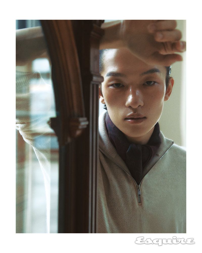 Portrait of an Asian adult male model with a buzzcut, wearing a sweater and scarf, leaning against a dark wood column indoors.