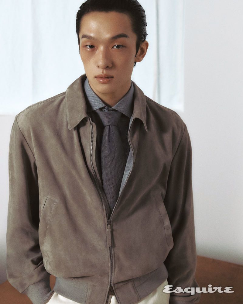 Adult male poses indoors wearing a grey button-up shirt, grey tie, taupe jacket, and white pants in a portrait fashion editorial.
