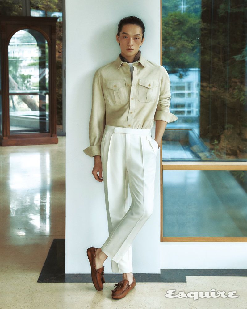 Portrait of a slender adult male in a beige shirt and white pleated pants, leaning against a wall in a bright, modern indoor space.