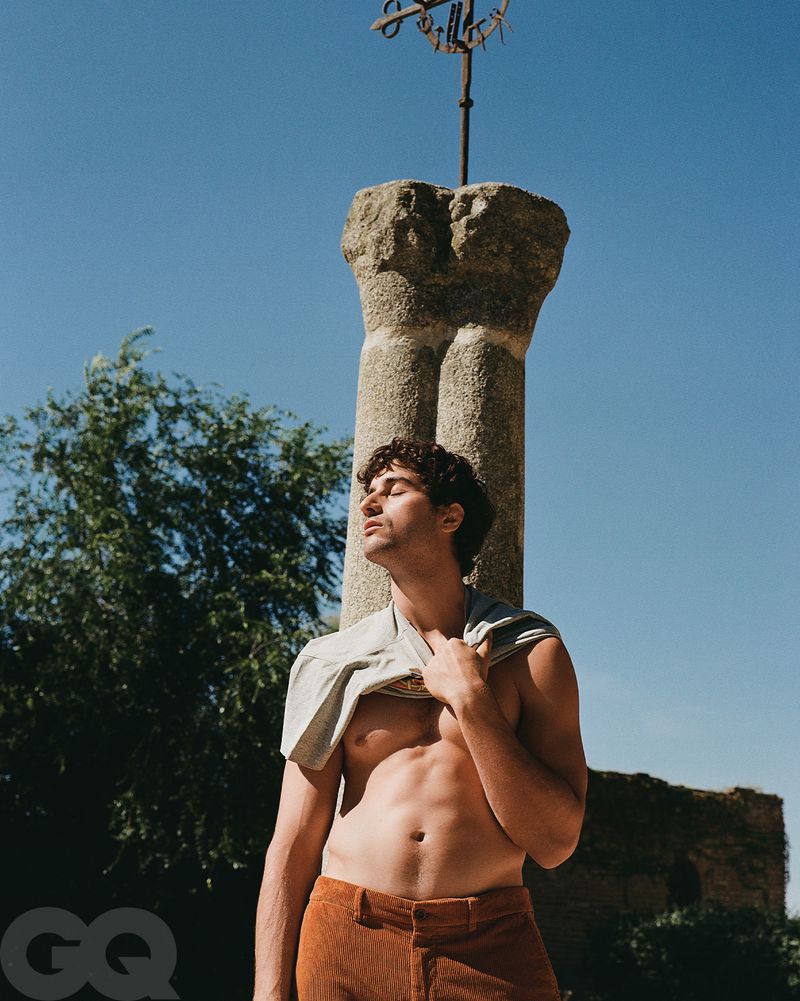 Man with exposed torso in outdoor setting, wearing corduroy trousers and a top around neck, near a stone pillar under a clear sky.