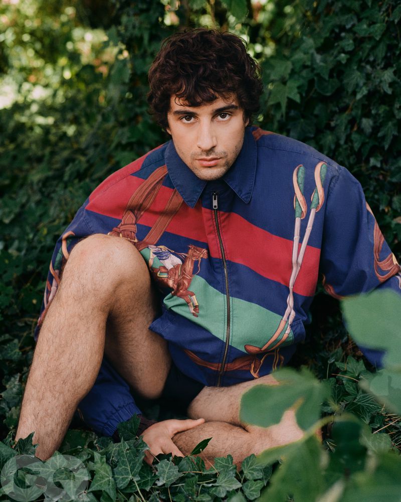Young adult man with curly hair and patterned jacket poses in natural setting surrounded by green foliage in a medium shot.