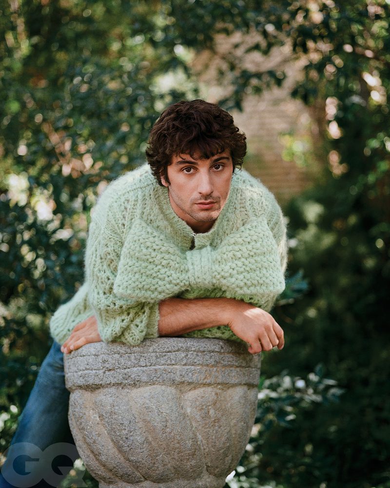 Portrait of a young adult male in a textured green sweater, leaning on a stone planter in an outdoor garden setting, medium shot.