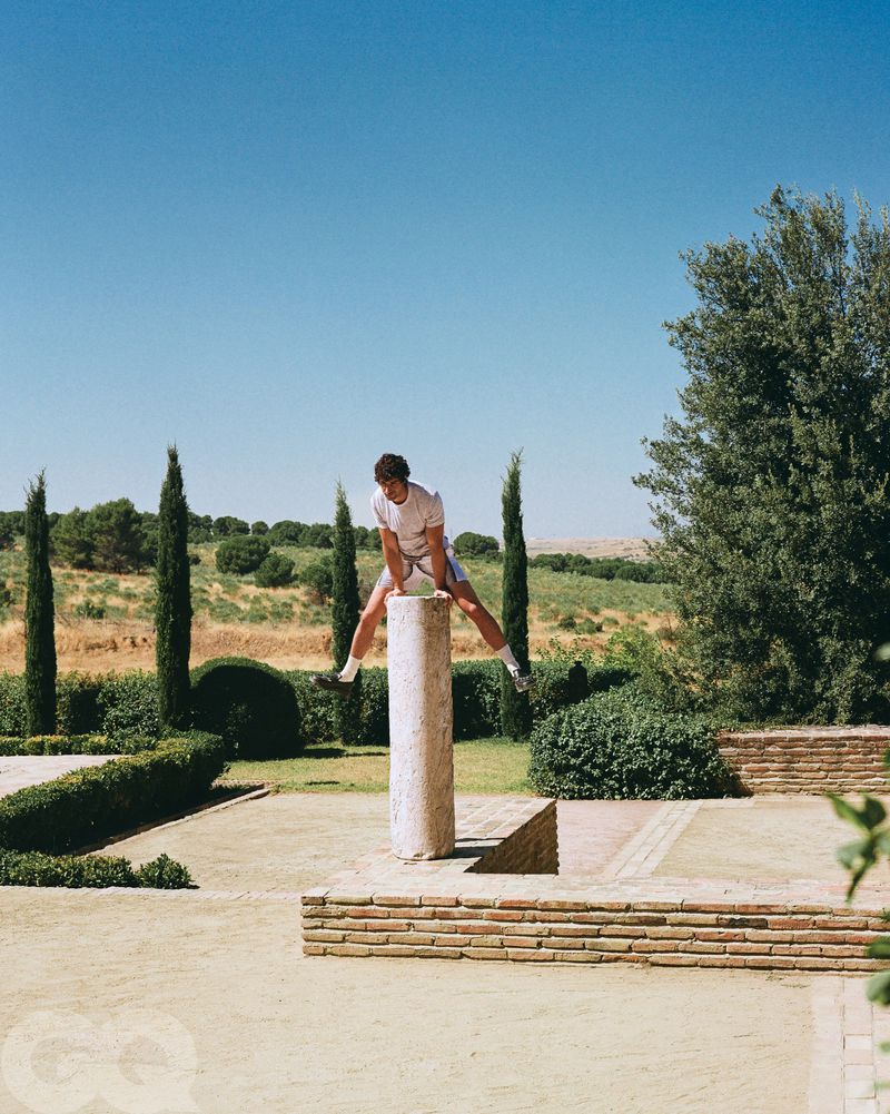 A man wearing a T-shirt and shorts is balancing on top of a stone column outdoors on a sunny day in a well-maintained grassy area.