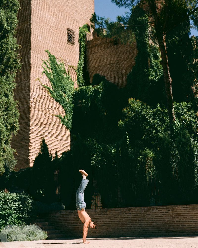 Shirtless adult male in jeans does a handstand on stone ground outside of brick building and green vegetation on a sunny day.