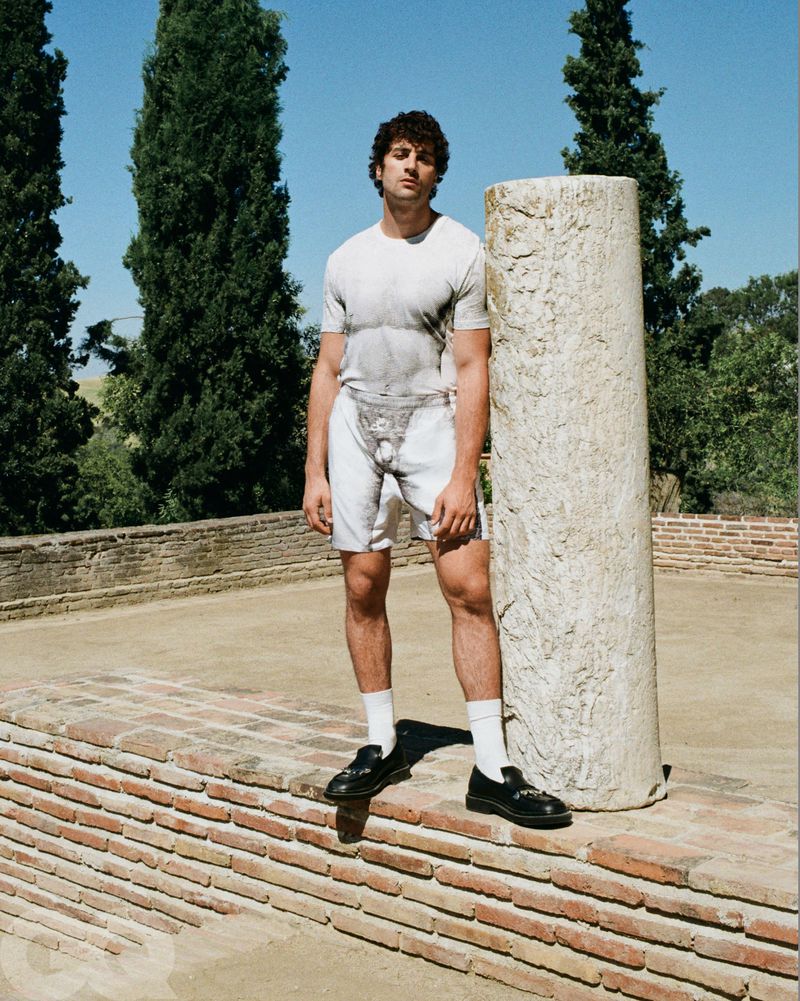 Young adult male poses on brick ledge next to stone pillar, wearing graphic t-shirt, shorts, socks, and loafers, with trees in background.