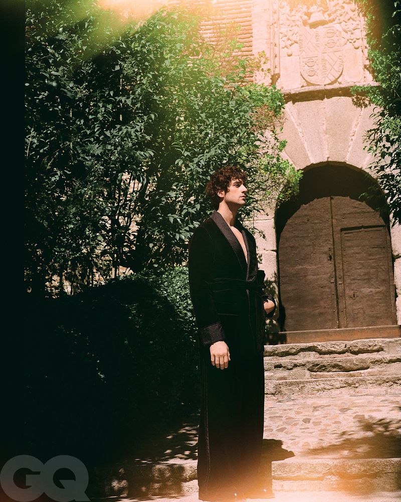 Man poses outdoors in a black robe on stone steps near a stone archway and building, with green foliage visible in the background.