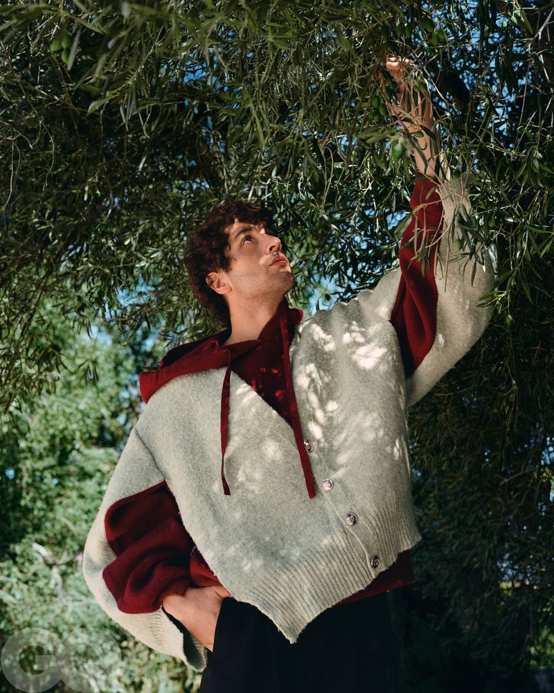 Man in hooded sweater stands under tree with green leaves, looking up with hand raised, photographed from a low angle in natural light.