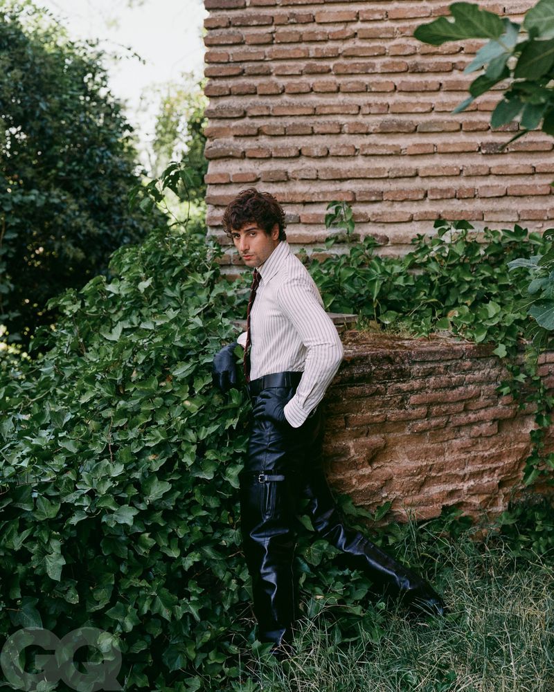 Male model poses outdoors against a brick wall and greenery, wearing a striped shirt, black tie, and glossy black pants for a fashion shoot.