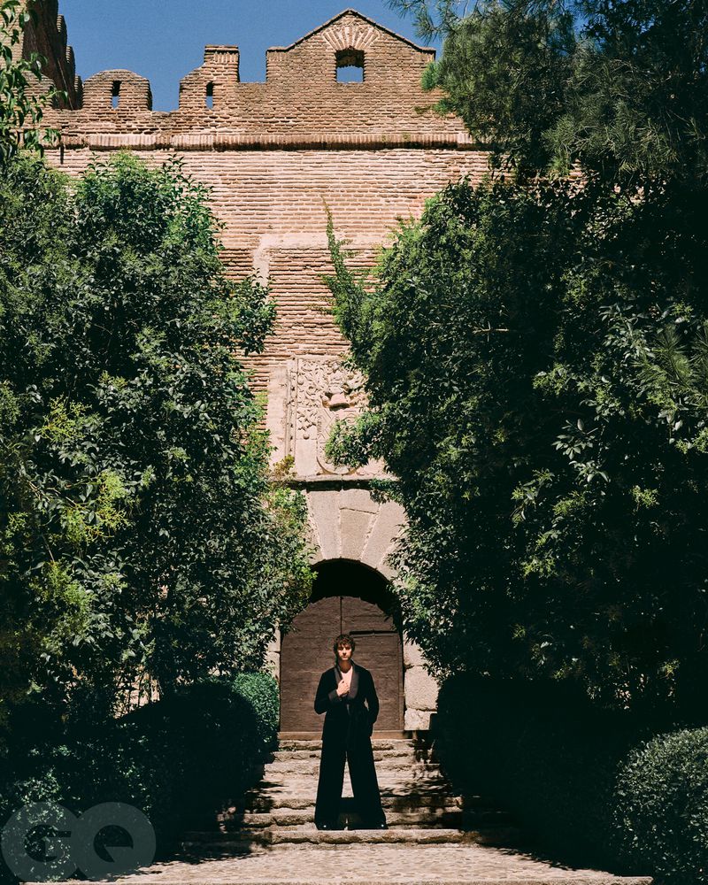 Adult man in black suit posing in front of a stone building and green trees on an overcast day. Fashion editorial image.