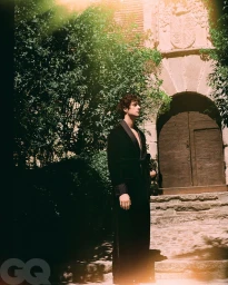 Agustin Della Corte stands in profile wearing a long black velvet belted robe outside a historic stone building with an arched doorway.