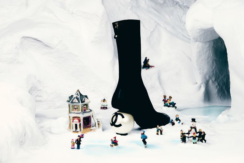 Chanel-branded sock on a logo-printed ball, surrounded by miniature figurines in a wintery, snow-covered landscape with a toy building.
