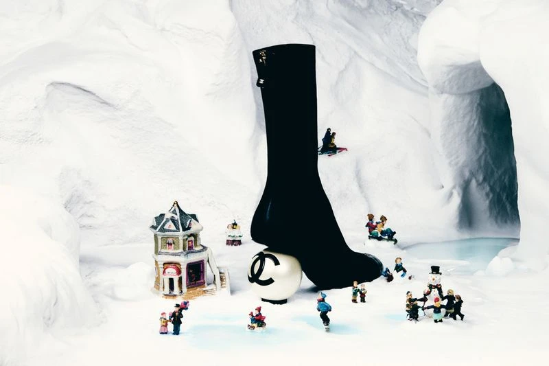 Chanel-branded sock on a logo-printed ball, surrounded by miniature figurines in a wintery, snow-covered landscape with a toy building.