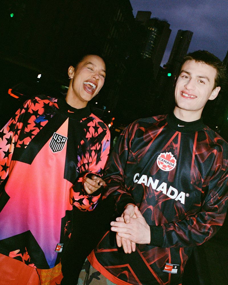 Two young adults in sports jerseys (USA, Canada) pose in a city street at night, sharing smiles and a sense of camaraderie.