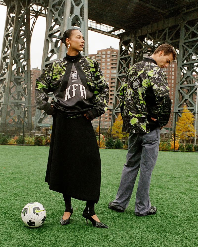 Two models in fashion apparel on a green field with a soccer ball, wearing patterned jackets and skirts, with a bridge backdrop.