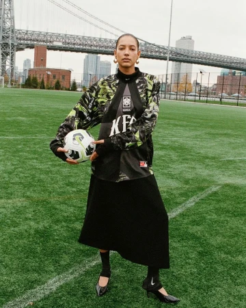 A woman in a streetwear-inspired outfit poses with a soccer ball on a green sports field, featuring a bridge and cityscape in the background.