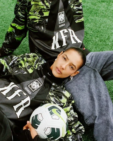 Woman in soccer jersey poses with soccer ball on grass, wearing jeans and hoop earrings with another person in the background.