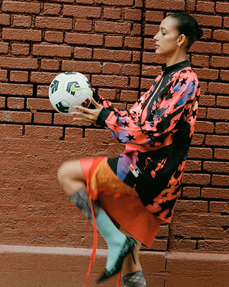 Adult female in athletic clothing balances a soccer ball against a brick wall, wearing high heel shoes and a patterned top.