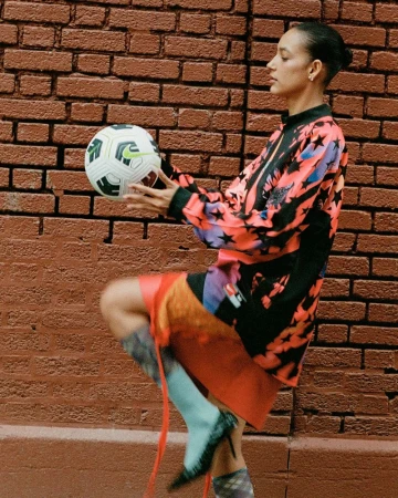 Adult female in athletic clothing balances a soccer ball against a brick wall, wearing high heel shoes and a patterned top.