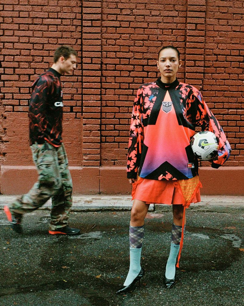 Two people stand in front of a brick wall; a woman holds a soccer ball and wears an athletic USA soccer jersey. A man walks by.