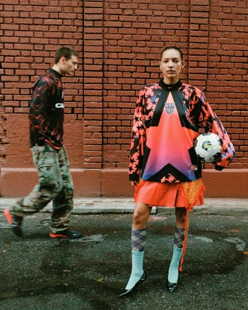 Two people stand in front of a brick wall; a woman holds a soccer ball and wears an athletic USA soccer jersey. A man walks by.