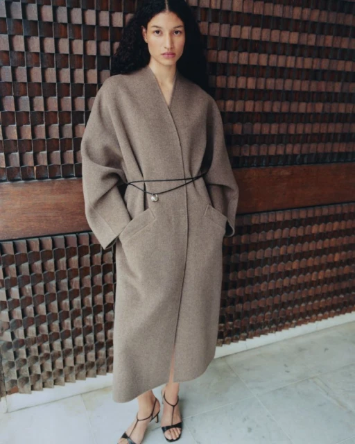 A woman wearing a long, brown coat with a black belt and black high heels, standing against a textured wall. Fashion editorial.
