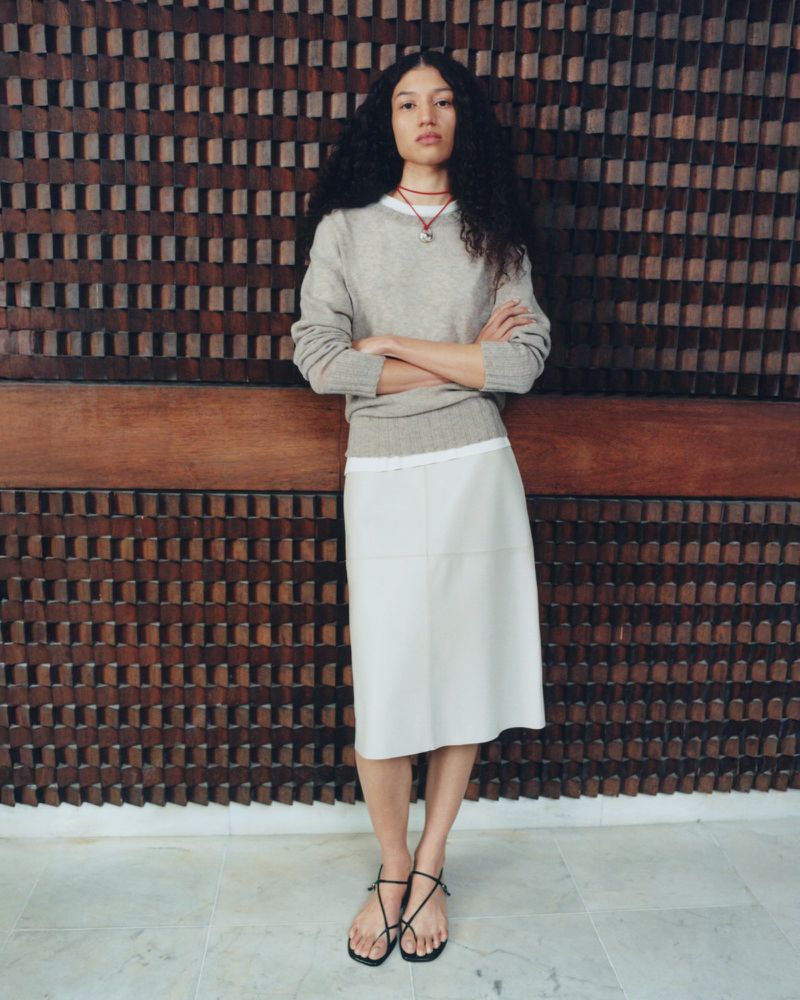 A long-haired woman poses indoors, wearing a beige sweater, white skirt, and strappy sandals, against a textured wooden wall.
