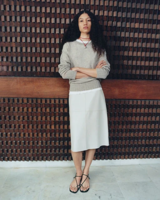 A long-haired woman poses indoors, wearing a beige sweater, white skirt, and strappy sandals, against a textured wooden wall.