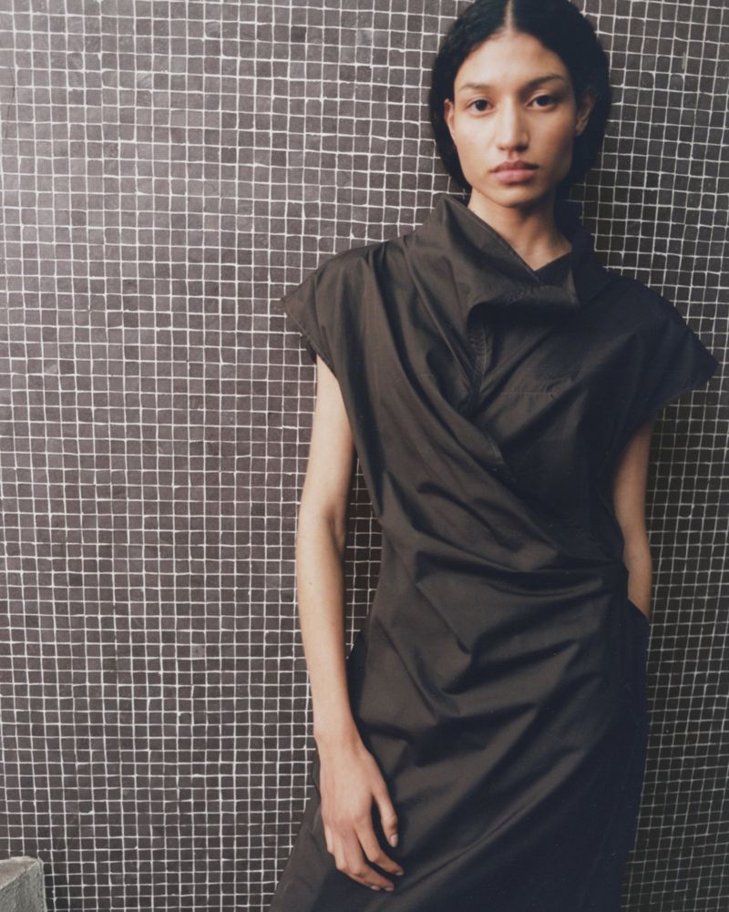 A fashion portrait of an adult female model in a dark dress with a unique neckline, posing against a square tiled wall indoors.