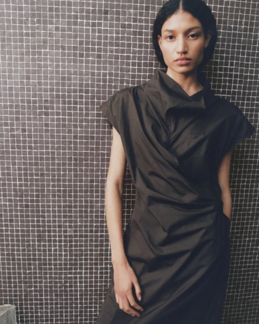 A fashion portrait of an adult female model in a dark dress with a unique neckline, posing against a square tiled wall indoors.