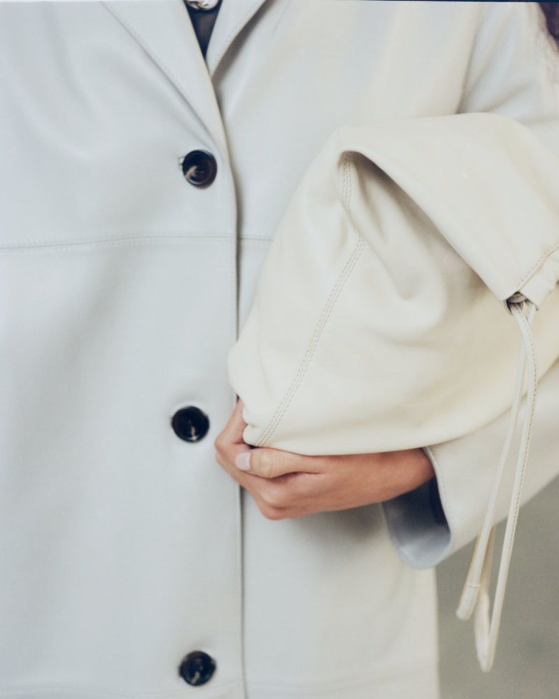 Fashion shot featuring an adult wearing a white jacket with dark buttons, holding a large, light-colored leather clutch bag.