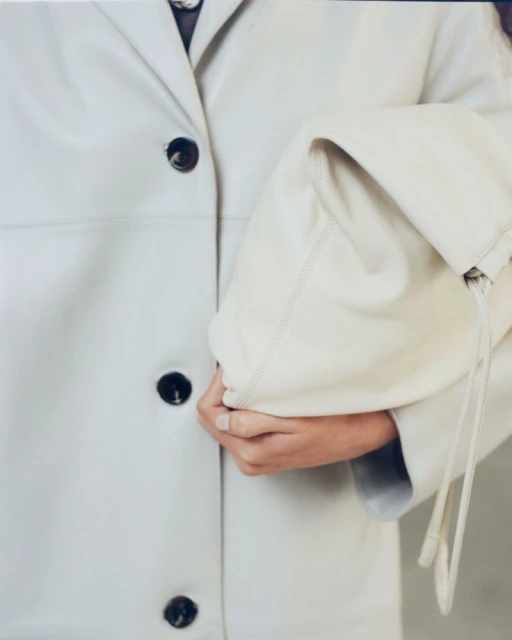 Fashion shot featuring an adult wearing a white jacket with dark buttons, holding a large, light-colored leather clutch bag.