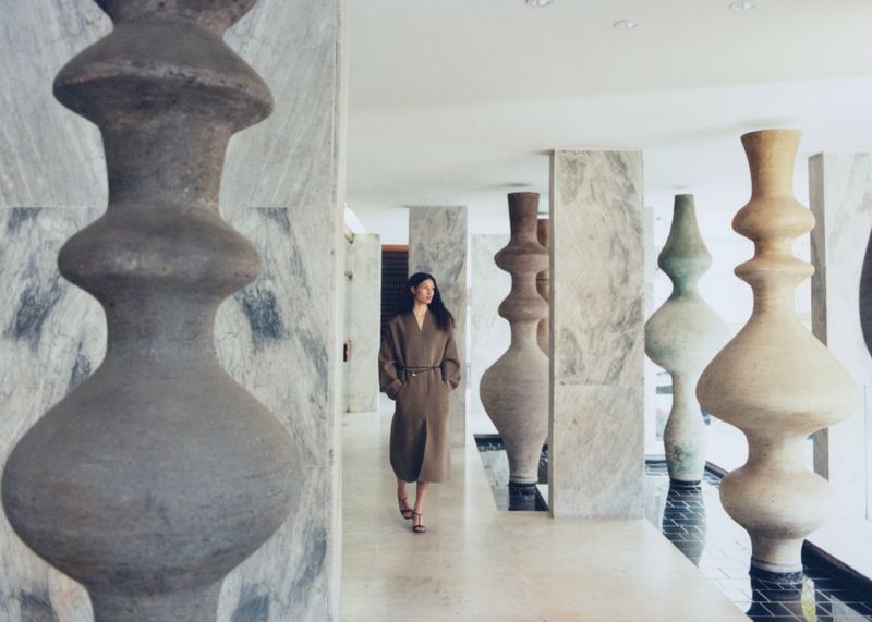 Woman in brown coat walking through modern corridor with abstract art installations and marble walls, bathed in soft, diffused lighting.
