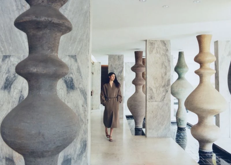 Woman in brown coat walking through modern corridor with abstract art installations and marble walls, bathed in soft, diffused lighting.